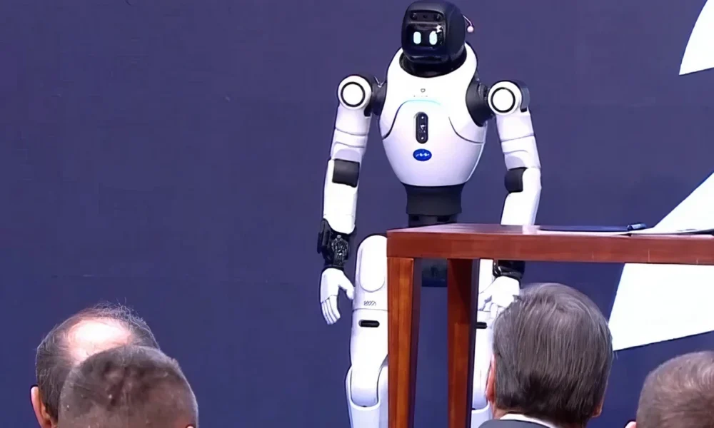 Egy emberszabású robot köszönti Aleksandar Vučić szerb elnököt (szürke öltönyben) és a mellette ülő Đuro Macut miniszterelnököt, akit kopaszodó fejéről lehet felismerni