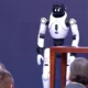 Egy emberszabású robot köszönti Aleksandar Vučić szerb elnököt (szürke öltönyben) és a mellette ülő Đuro Macut miniszterelnököt, akit kopaszodó fejéről lehet felismerni