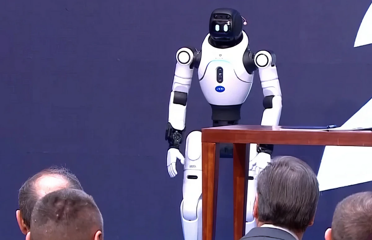 Egy emberszabású robot köszönti Aleksandar Vučić szerb elnököt (szürke öltönyben) és a mellette ülő Đuro Macut miniszterelnököt, akit kopaszodó fejéről lehet felismerni